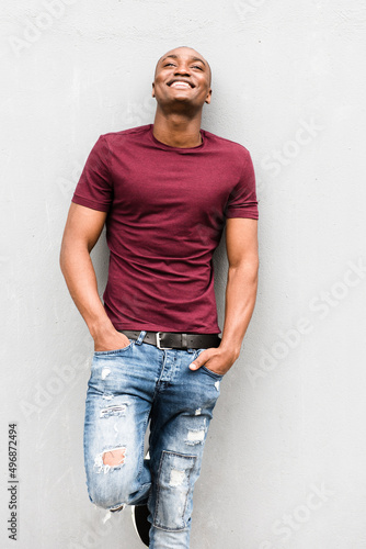 happy young man leaning against gray wall with hands in pocket and looking up