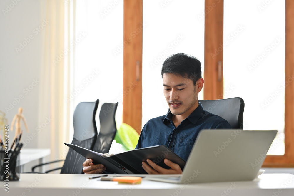 A portrait of a young Asian man sitting in the office holding and looking at the report, data and documents, for business and technology concept. A portrait of a young Asian man sitting in the office holding and looking at the report, data and documents, for business and technology concept.