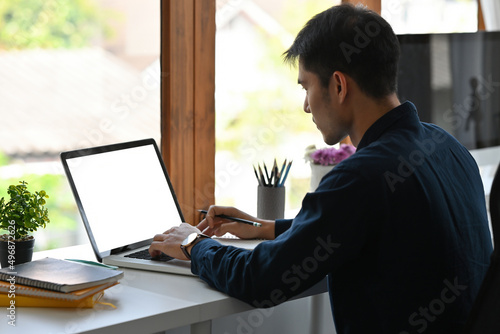 A portrait of a handsome Asian businessman sitting in the office and working on a laptop with white blank screen, for business and technology concept.