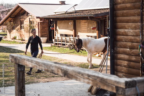 Smiling Caucasian woman walking with the pony among stables on countryside ranch