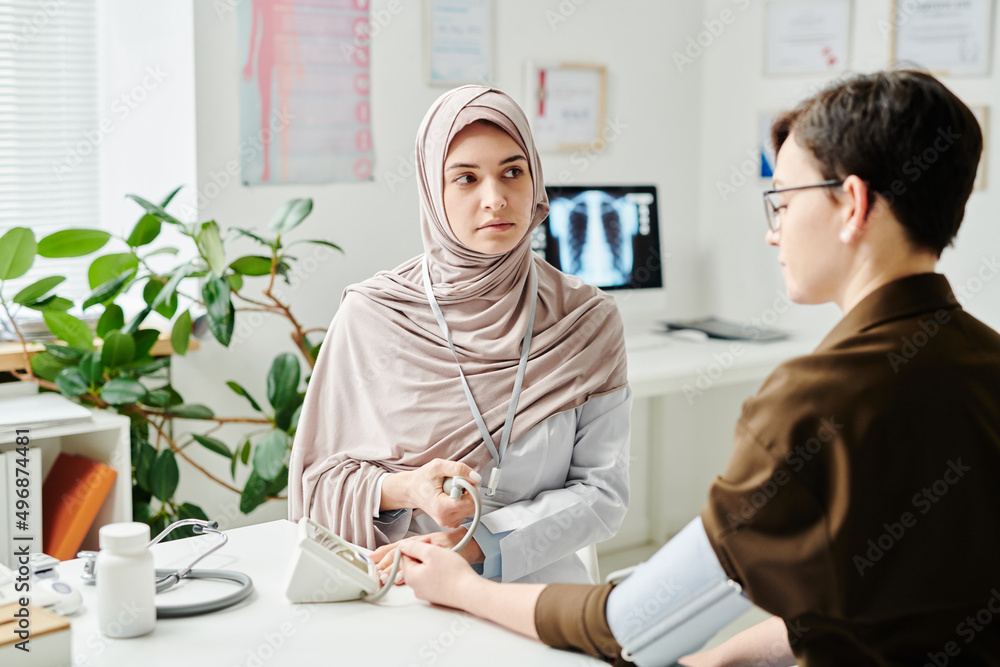 Obraz premium Young serious Muslim doctor measuring blood pressure of sick female patient sitting in front of her by desks in medical office