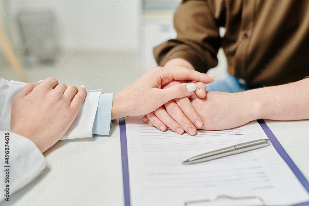 Obraz premium Hand of doctor in whitecoat on those of sick female patient sitting by desks in front of her during support after announcing diagnose
