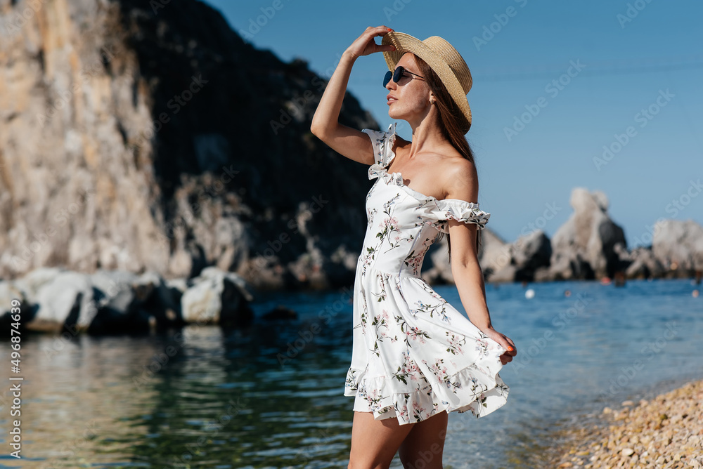 A beautiful young woman in a hat, glasses and a light dress is walking along the ocean shore against the background of huge rocks on a sunny day. Tourism and tourist trips.