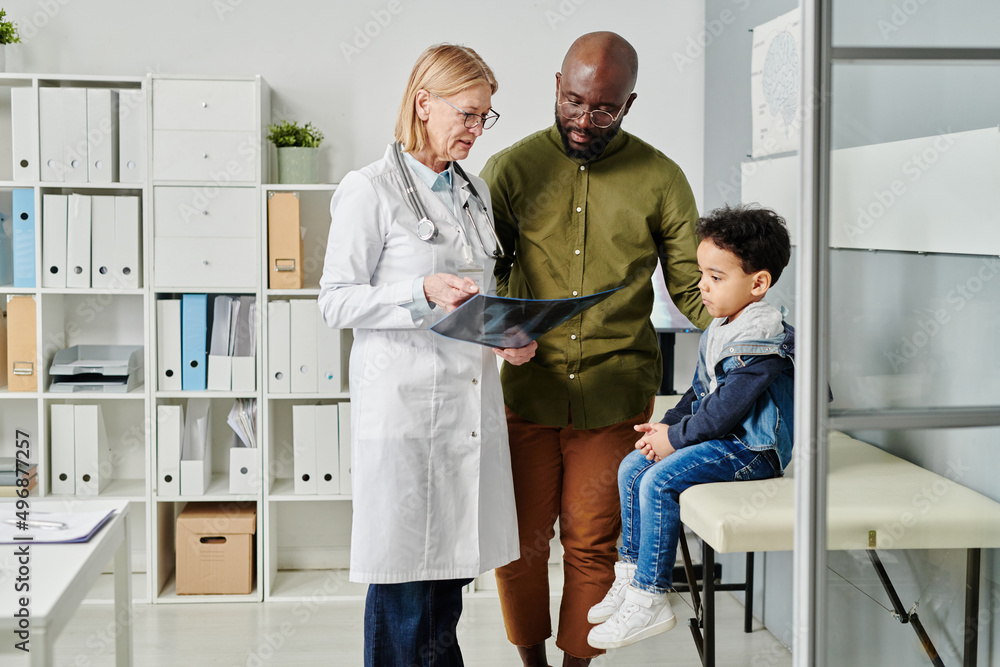 Obraz premium Young African American man and his cute little son listening to explanation of mature female radiologist with x-ray image