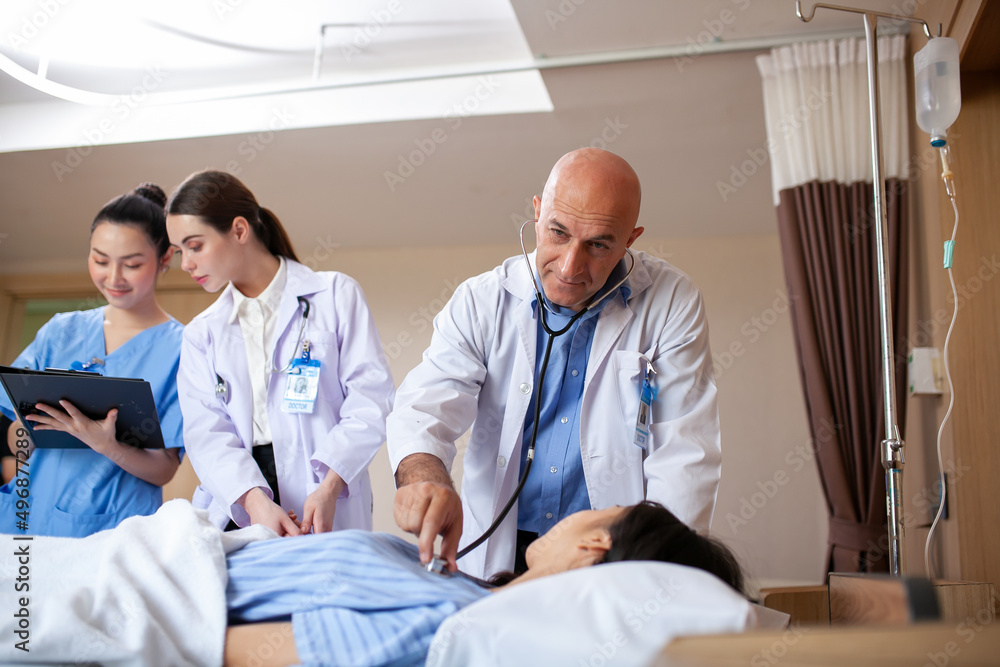 Obraz premium Group of doctor visiting ward have woman patient and check her with stethoscope on bed. Woman patient lying on hospital bed while doctor giving her checkup.