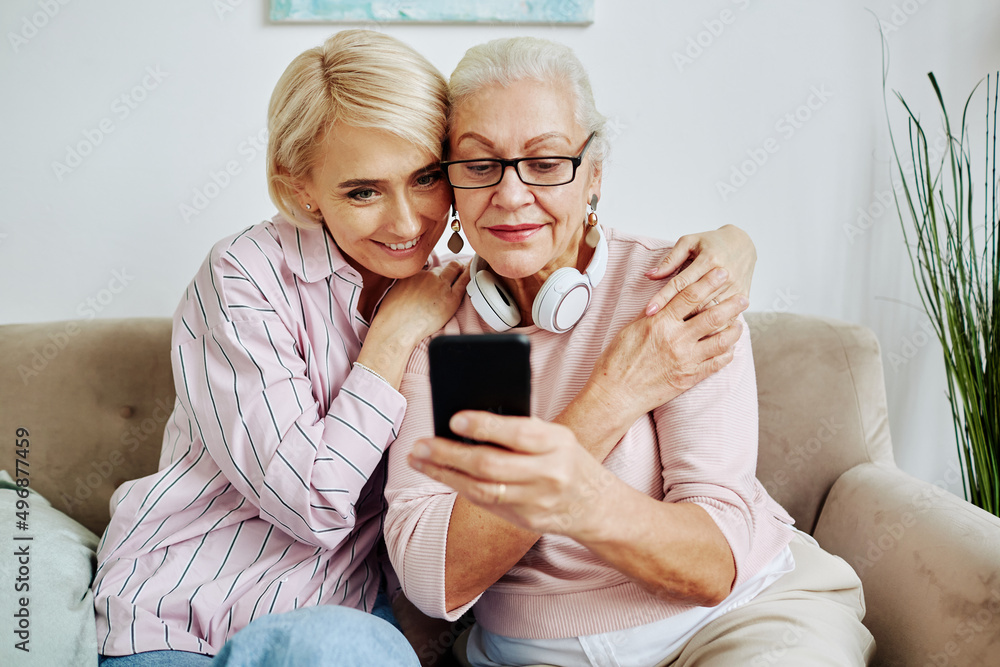 Portrait of modern senior woman using smartphone device at home with smiling young daughter embracing her