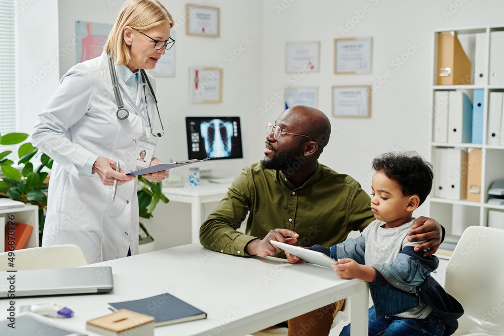 Mature female doctor in whitecoat standing by workplace and consulting young African American man with little son in clinics Mature female doctor in whitecoat standing by workplace and consulting young African American man with little son in clinics