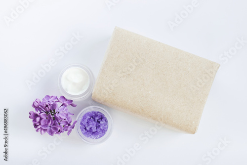 Soap in craft paper, sugar scrub, cream with lilac flowers on white background. Mockup and copy space. Spa treatments for home care. Home rejuvenation and moisturizing