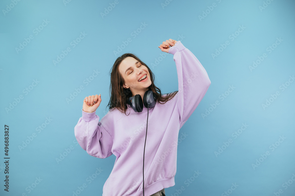Portrait of a joyful lady with braces on her teeth and headphones on her neck dancing with a smile on her face on a blue background. Portrait of a joyful lady with braces on her teeth and headphones on her neck dancing with a smile on her face on a blue background.