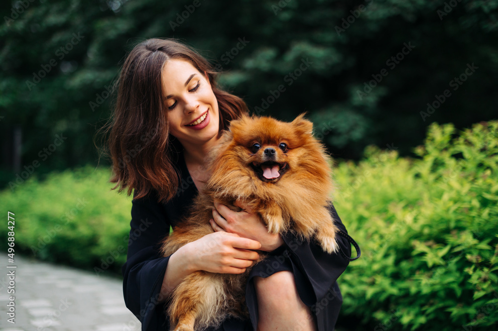 Portrait of an attractive brunette woman with a cute Pomeranian Spitz dog in her arms, looking at a pet and smiling.