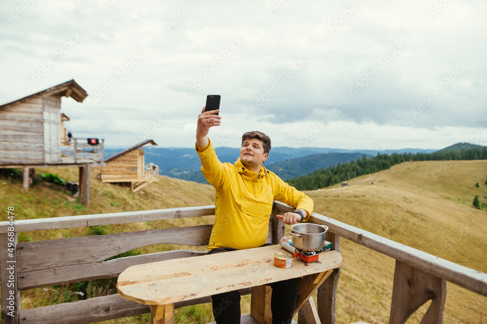 Positive man is sitting on a bungalow terrace at a table in the mountains, preparing food and using a smartphone with a cheerful face. Positive man is sitting on a bungalow terrace at a table in the mountains, preparing food and using a smartphone with a cheerful face.