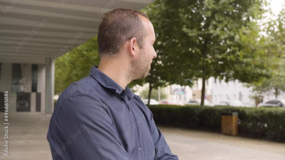 A Caucasian man waves at the camera with a smile by an office building in an urban area - a street in the blurry background A Caucasian man waves at the camera with a smile by an office building in an urban area - a street in the blurry background