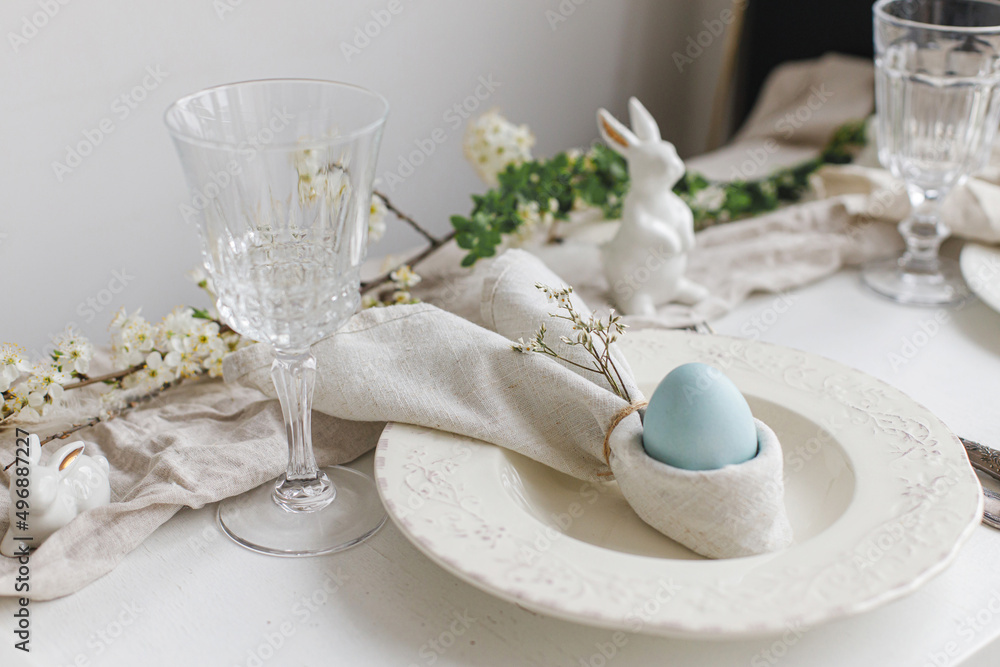 Easter egg in bunny napkin on plate with cutlery, bunny, spring flowers on rustic white table. Stylish elegant Easter brunch table setting. Modern table arrangement and eco decoration Easter egg in bunny napkin on plate with cutlery, bunny, spring flowers on rustic white table. Stylish elegant Easter brunch table setting. Modern table arrangement and eco decoration
