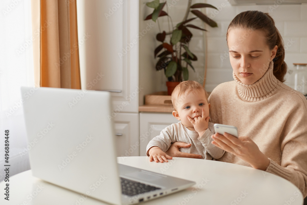 Indoor shot of Caucasian dark haired woman wearing beige sweater sitting at table with baby girl in hands and using cell phone, holding mobile pone in hands. Indoor shot of Caucasian dark haired woman wearing beige sweater sitting at table with baby girl in hands and using cell phone, holding mobile pone in hands.