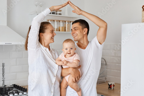 Smiling positive family posing looking at each other with happy facile expression, holding baby daughter in hands, mother and father making roof figure with hands arms over heads.