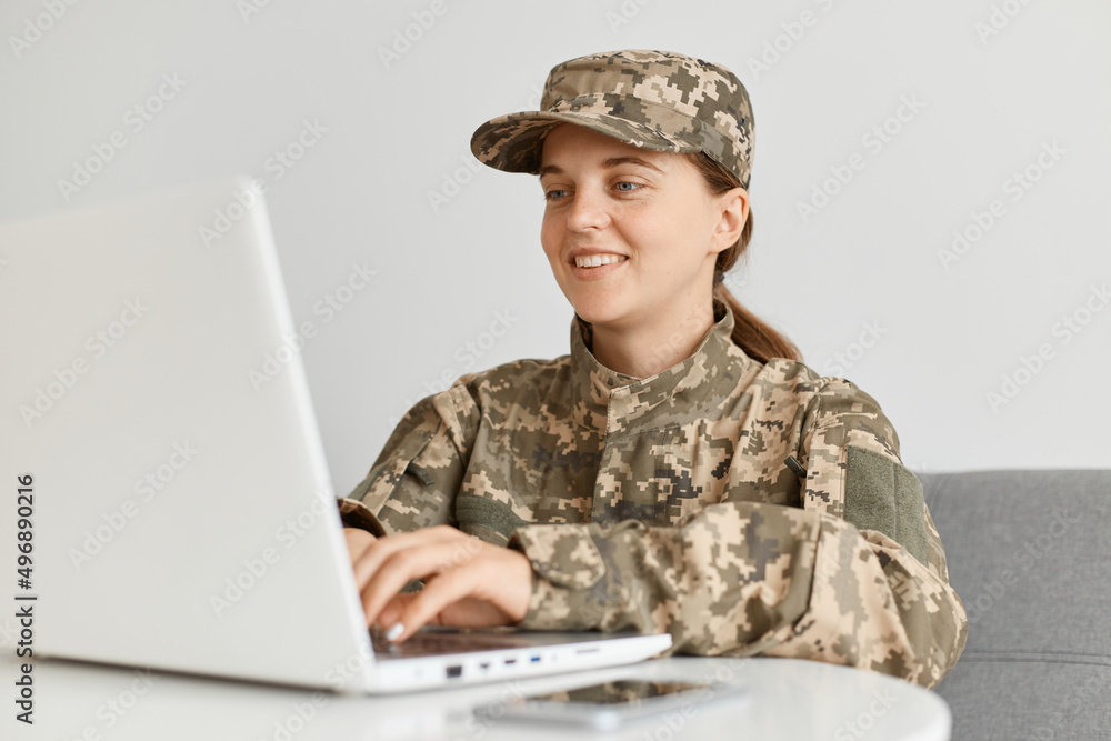 Fototapeta premium Portrait of smiling young adult beautiful woman wearing camouflage uniform and cap, working on portable computer, being in good mood, expressing happiness.