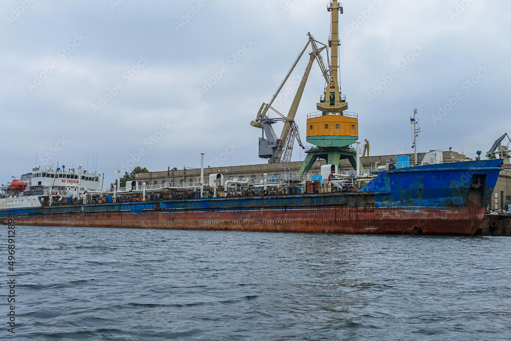 An old, rusty oil or gas ship stands in port for loading or unloading. In the background are port cranes and a cloudy sky.