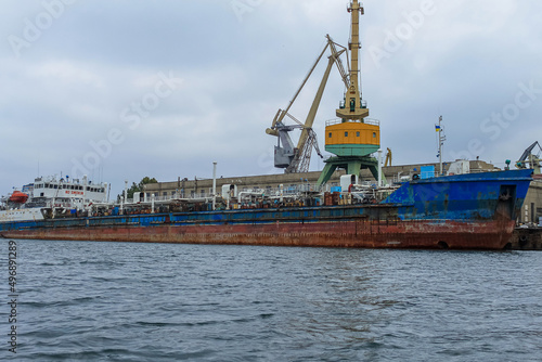An old, rusty oil or gas ship stands in port for loading or unloading. In the background are port cranes and a cloudy sky.