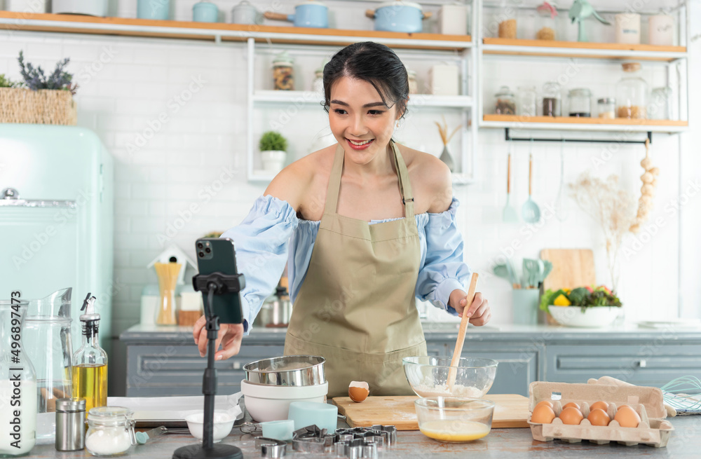 Asian woman food blogger cooking cake in front of smartphone camera while recording vlog video and live streaming at home in kitchen. Asian woman food blogger cooking cake in front of smartphone camera while recording vlog video and live streaming at home in kitchen.