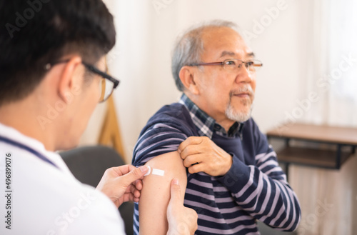Safe vaccination for old people. Elder man in medical getting flu or Covid-19 vaccine sitting on sofa at home. Asian Doctor or nurse giving flu or Covid-19 shot to senior.
