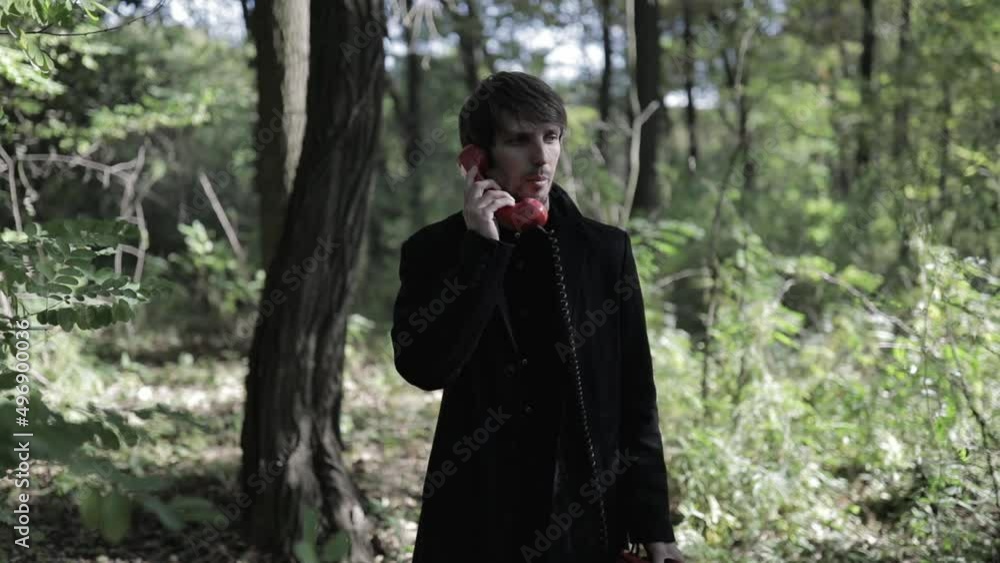 A man in a black coat is talking on a red vintage telephone in the forest. creative shooting A man in a black coat is talking on a red vintage telephone in the forest. creative shooting