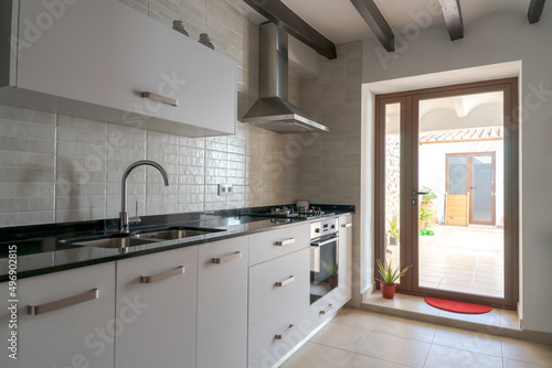 White kitchen with natural light from the yard. White furniture with black marble. Ceiling with wooden beams. Gas hob and stainless steel extractor fan.