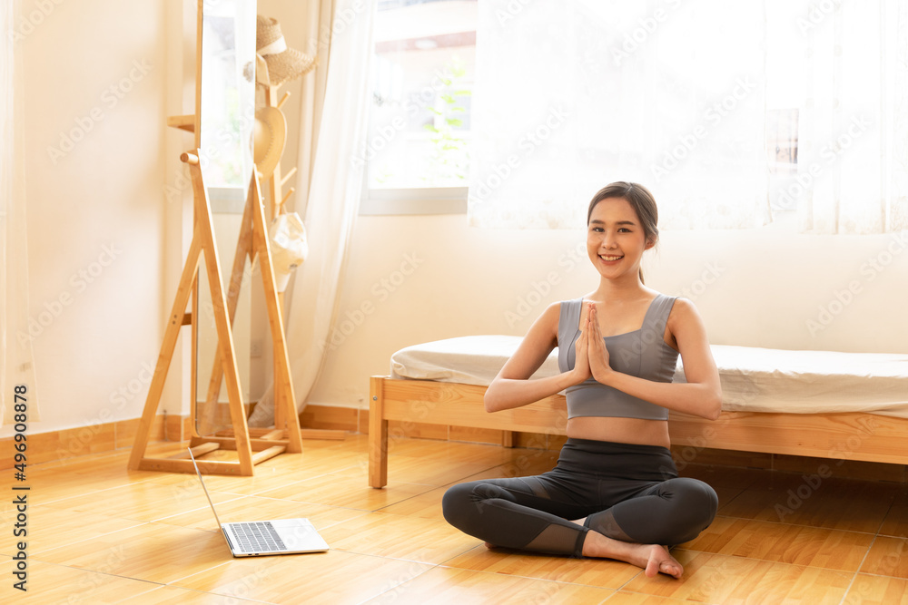 Young Asian woman is exercising yoga at home. Fitness, workout, healthy living and diet concept. Female watching videos online on laptop computer in bedroom.
