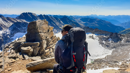 Hiking man with big backpack sitting. Scenic view on Hoher Sonnblick glacier, High Tauern mountains in Carinthia, Salzburg, Austria, Europe, Alps. Goldbergkees in Hohe Tauern National Park, sunny day photo