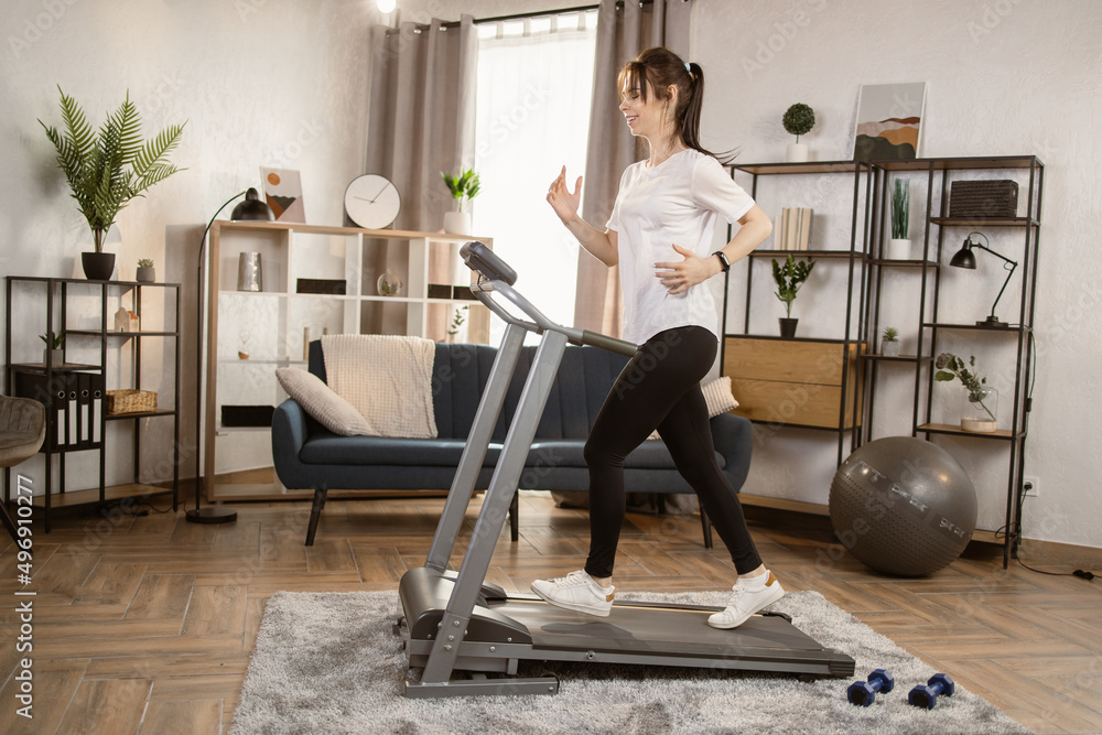 Attractive young sports caucasian woman running on treadmill. Doing fitness exercise. Athletic and muscular woman actively workout in the modern apartment. Sports woman workout at home. Side View