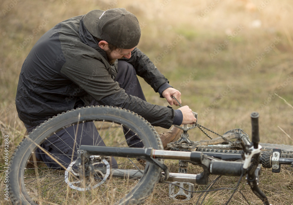 a young cyclist is sitting on the grass and repairing a loose chain on a mountain bike..