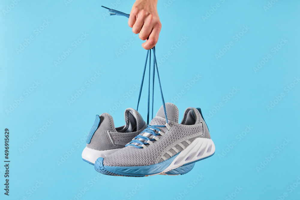 Hand holds hanging gray running sneakers by the laces on blue pastel