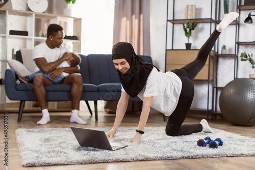 Fototapeta Naklejka Na Ścianę i Meble -  Fit muslim arabic woman doing yoga plank and watching online tutorials on laptop while training in living room and her african husband puts little son to sleep while sitting on the couch.