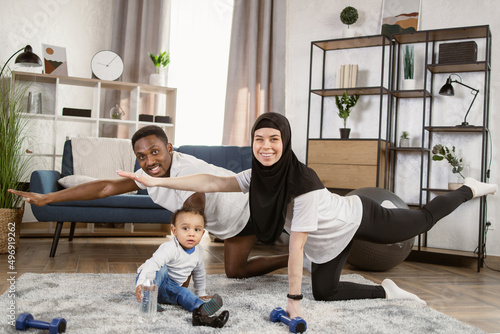 Young attractive family african man and muslim arabic woman practising yoga stretching workout at home looking for their cute little son playing on the carpet, healthy lifestyle body care.