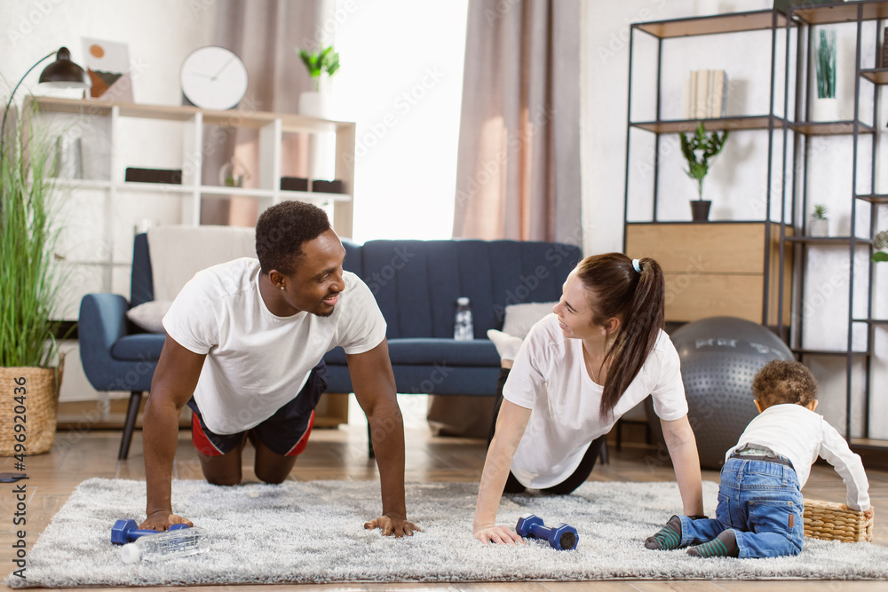 Young attractive family african man and woman doing push up exercises workout at home looking for their cute little son playing on the carpet, healthy lifestyle body care. Young attractive family african man and woman doing push up exercises workout at home looking for their cute little son playing on the carpet, healthy lifestyle body care.