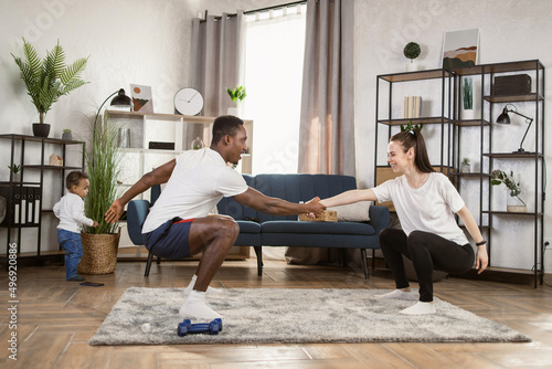 Young multinational multiracial couple doing squats holding hands training together at home, african man and caucasian woman working out together while their son is playing in the background.