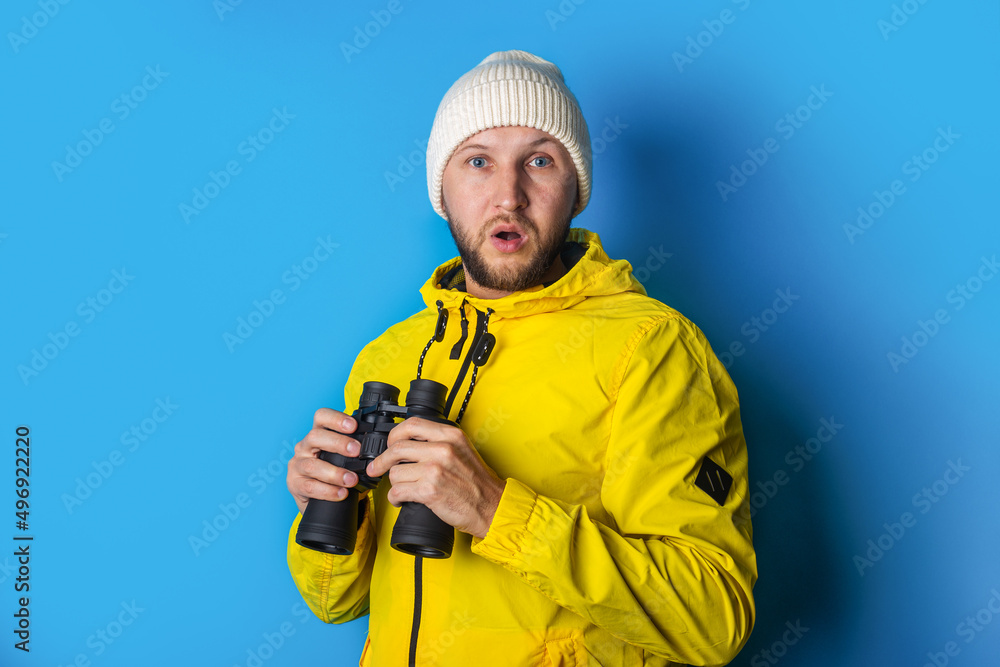 surprised shocked young man in a yellow jacket with binoculars on a blue background