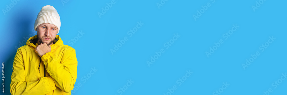 Pensive young man in a yellow jacket holding his hand at his chin on a blue background. Banner Pensive young man in a yellow jacket holding his hand at his chin on a blue background. Banner