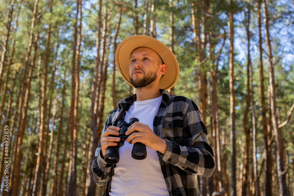 Man tourist in a hat and a plaid shirt looks through binoculars in the forest
