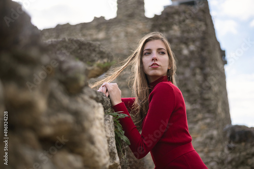 Young girl in the red dress stands near the ancient stone wall among the old fortress. © Dmitriy