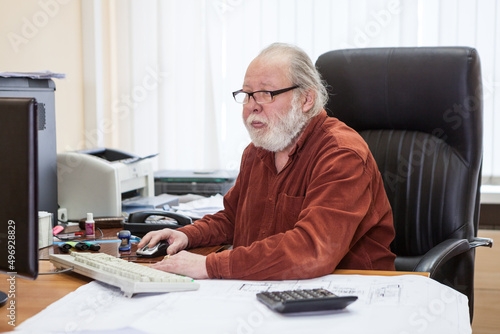 Concentrated bearded senior man using computer at desk in office room, engineer white collar worker