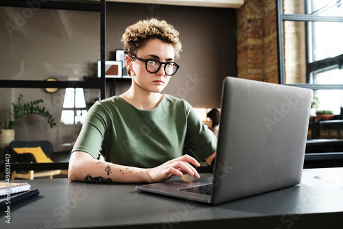 Young woman in eyeglasses concentrating on her online work on laptop at her workplace at office