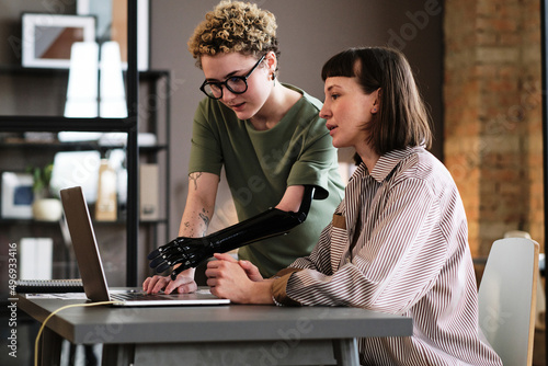 Young woman with prosthetic arm pointing at laptop and talking to her colleague during work at office photo