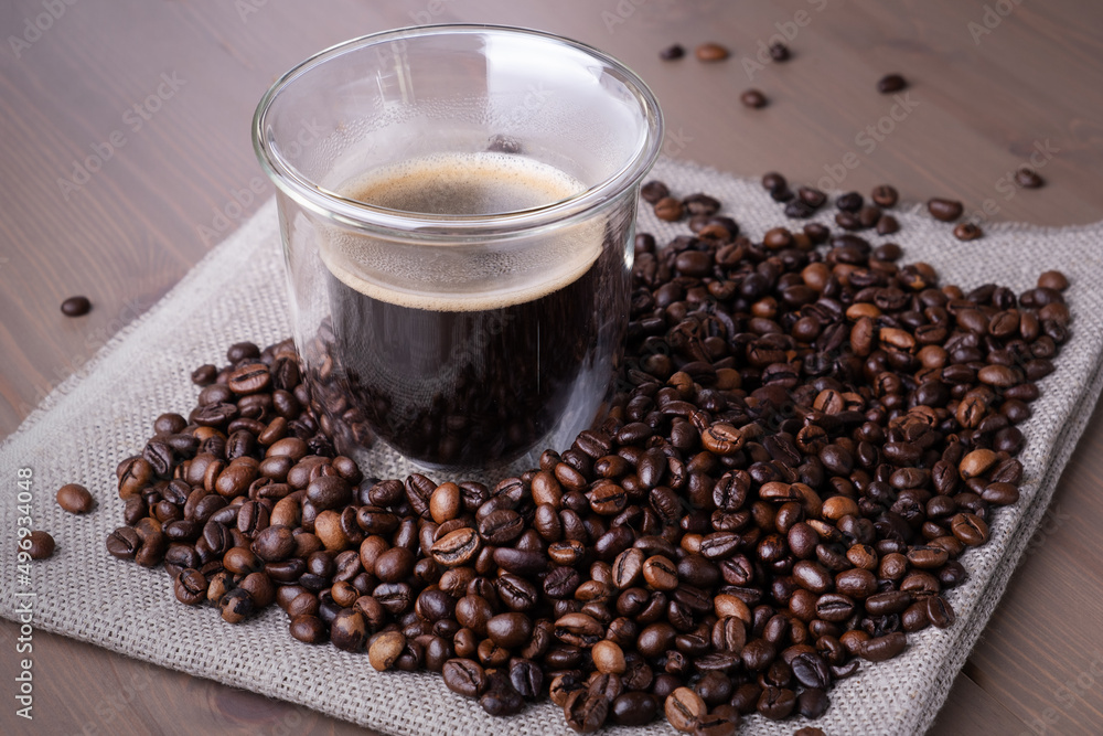 Fototapeta premium Close-up of glass cup with coffee and pile of roasted coffee beans on piece of burlap on wooden surface. Heat-resistant coffee glass with double walls on table. Selective focus.