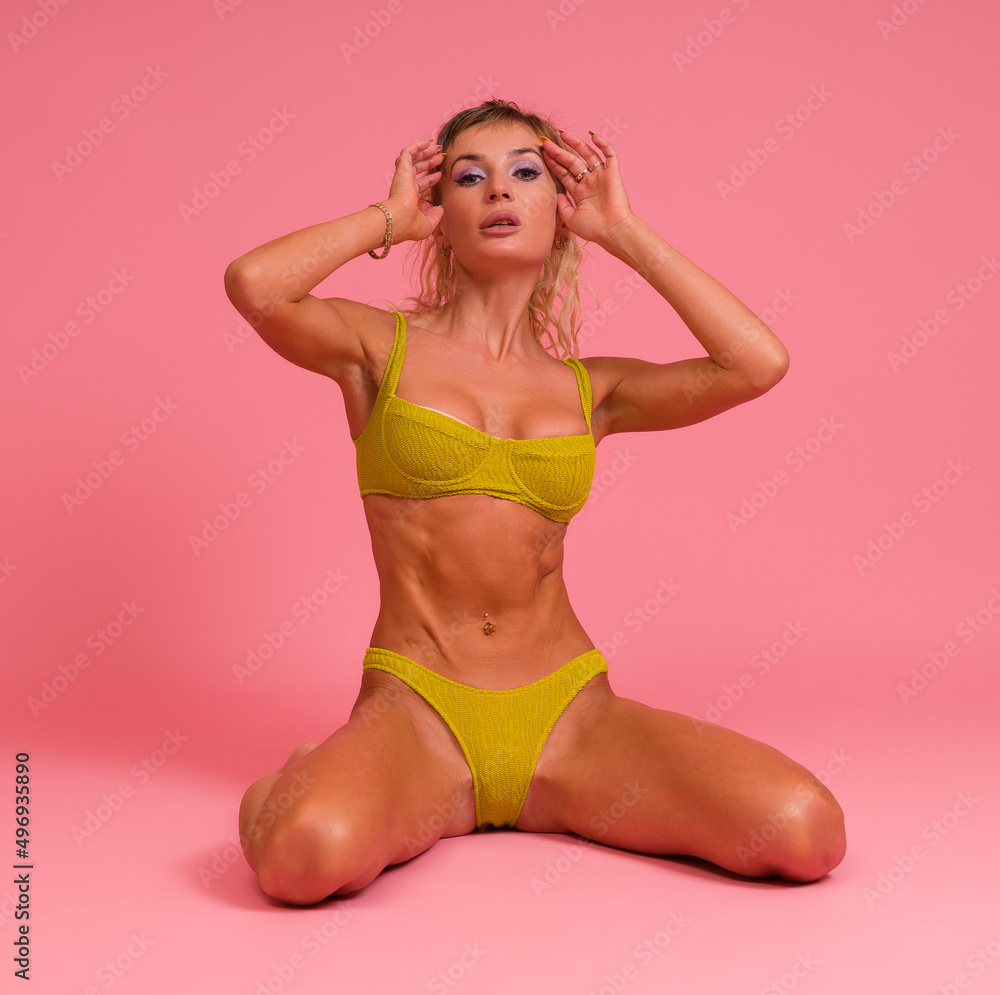 Naklejka premium Beautiful sexy woman in a yellow swimsuit posing sitting on her knees on a pink background.
