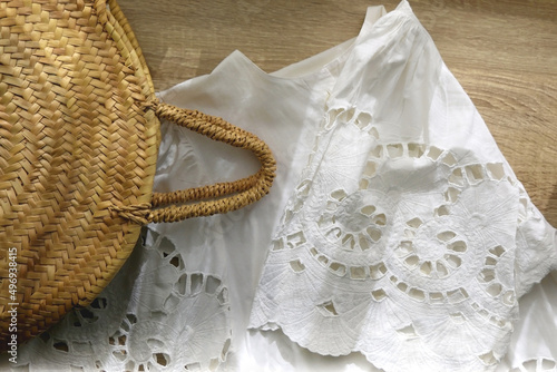 White linen lace blouse and round straw bag on wooden background. Flat lay.