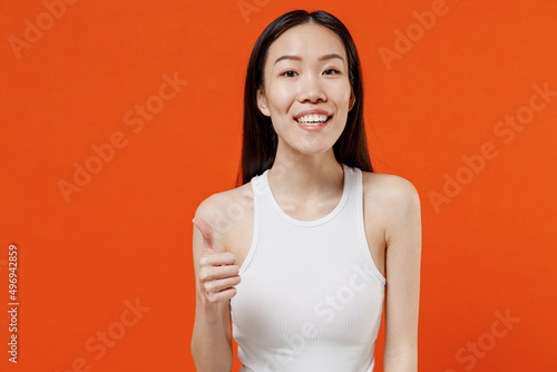 Overjoyed fancy vivid excited smiling jubilant exultant young woman of Asian ethnicity 20s year old in white tank top showing thumb up like gesture isolated on plain orange background studio portrait