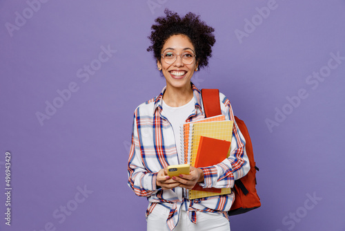 Young fun girl woman of African American ethnicity student in shirt backpack hold books use mobile cell phone isolated on plain purple background. Education in high school university college concept.