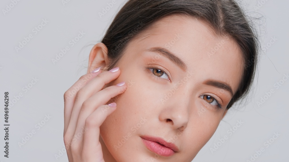 Extreme close-up beauty portrait of young dark-haired woman touches her face sliding fingers from a cheek down to her chin, looks at camera and smiles | Skincare product commercial concept