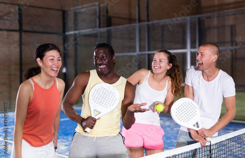 Happy laughing men and women of different nationalities in sportswear with rackets and balls in hands talking friendly near net on indoor padel court. © JackF