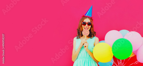 Happy smiling young woman in birthday hat with colorful balloons eating lollipop on pink background, blank copy space for advertising text